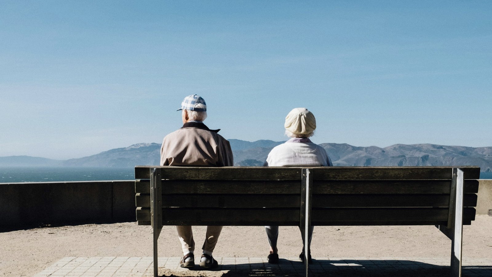 Two people sitting together on a bench, looking out across water toward distant hills.
